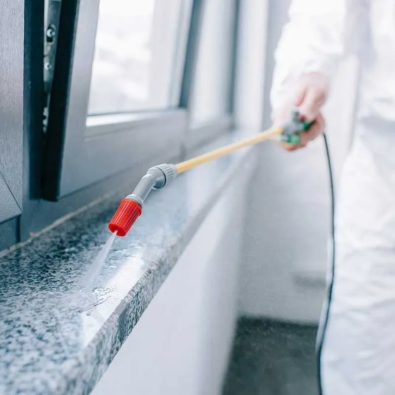 A person in a white protective suit uses a long-handled sprayer to apply liquid to a granite windowsill indoors.