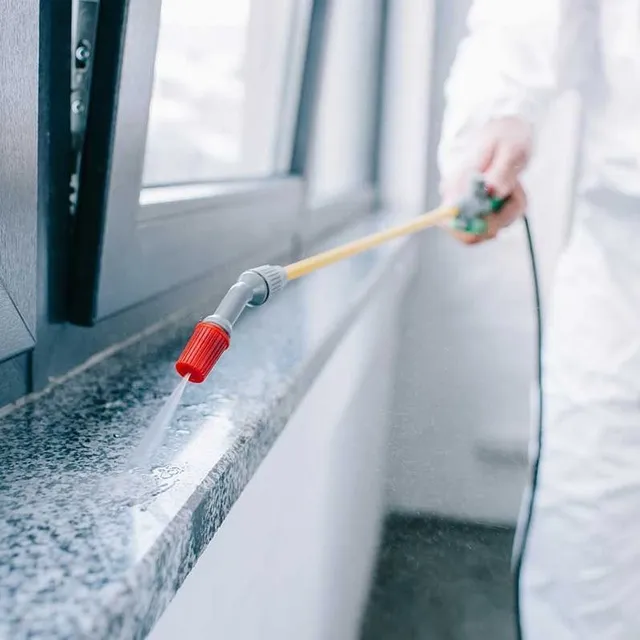 A person in a white protective suit uses a long-handled sprayer to apply liquid to a granite windowsill indoors.