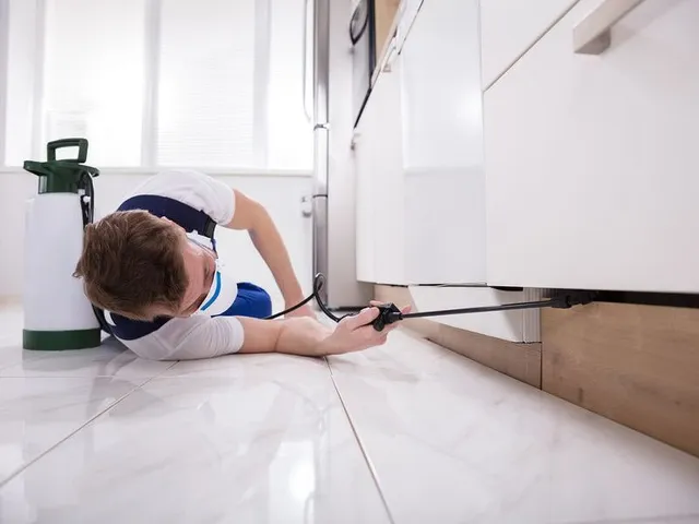 A service professional lying on a tiled floor, using a spray wand to apply pest control under kitchen cabinets.